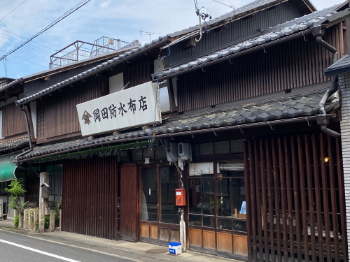 岡田防水布店と誇らしげに書かれた古い看板が掲げられた、味わい深い木造店舗の正面外観