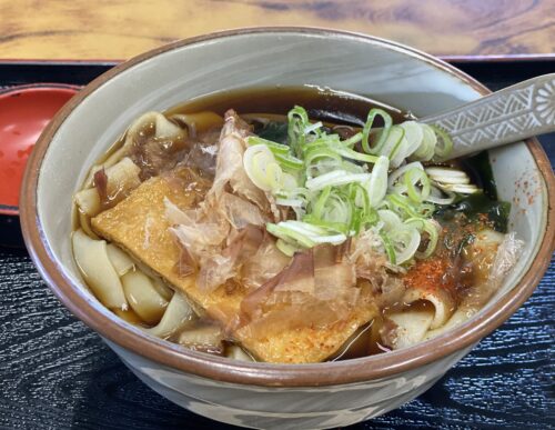 『名物きしめん（900円）』。醤油ベースの澄んだお出汁に、大ぶりの平打ちきしめん、お揚げ、蒲鉾、小松菜が乗り、たっぷりの鰹節がふわりと踊っています。刻みネギは小皿で別添えに。箸袋には名古屋城と金シャチのイラスト、箸置きを兼ねた「きしめん亭」のロゴがあります。